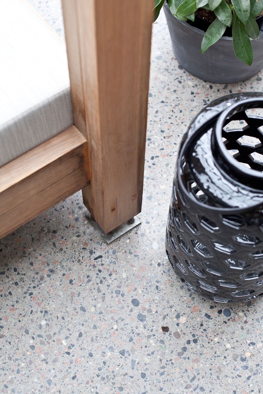 Nærbilde av trebenk med lys grå pute på terrazzo-gulv, sort perforert keramikklykt og grønn plante.