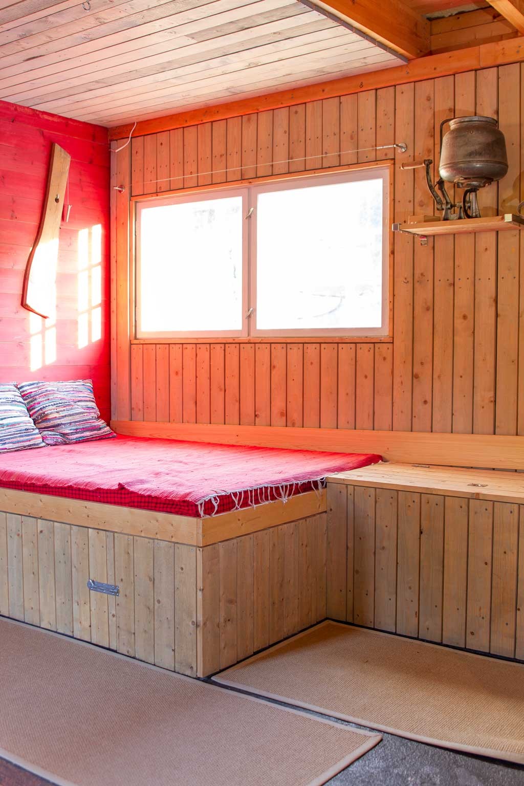 Lyst trepanelrom med innebygd benk, rødt teppe, stripete puter, vindu og metallkanne