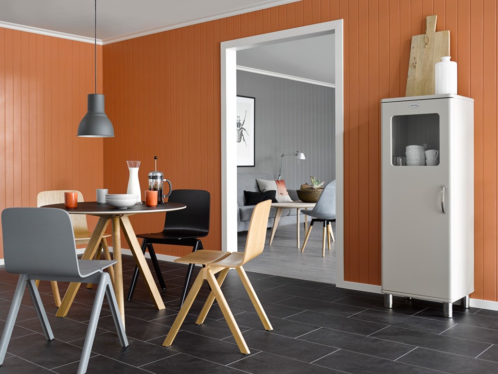 Moderne spisestue med rundt trebord, stoler mot oransje panelvegg og hvitt skap, grå stue bak