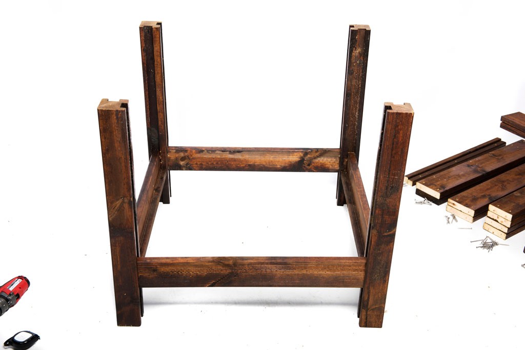 Opp-ned mørkebrunt trebordramme på hvit bakgrunn med løse planker, skruer og rød drill.