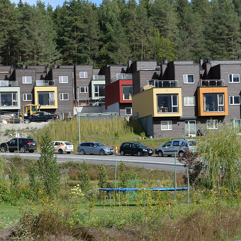 Mørkebrune rekkehus med fargesterke balkongbokser i gult, rødt og orange, foran vei og skog.