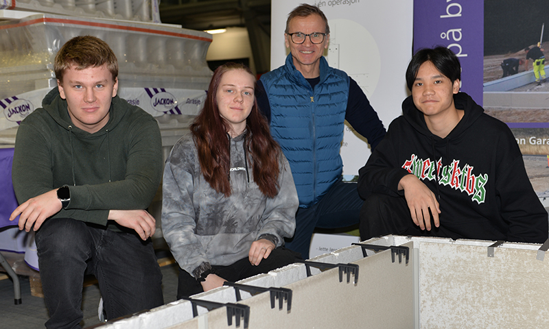 Fire personer ved hvite isolasjonsblokker og lilla-hvitt messebannere.