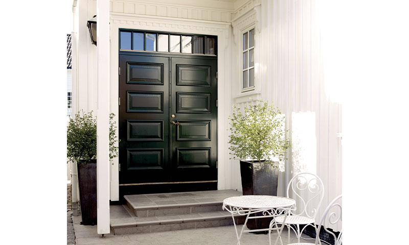 Inngang med svart dobbeltdør i hvit fasade, to potteplanter og hvitt hagebord.