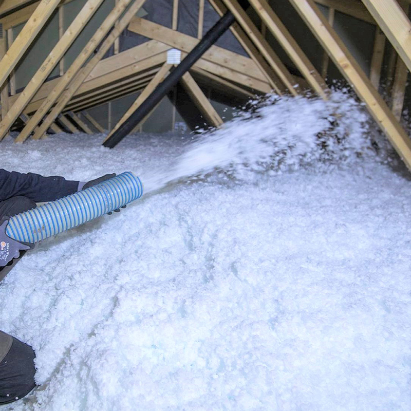 Loft med lyse trebjelker og blå slange som sprøyter hvit celluloseisolasjon