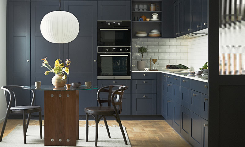 Moderne kjøkken med mørkeblå skap, hvit flis, glassbord med vase, trestoler og pendellampe