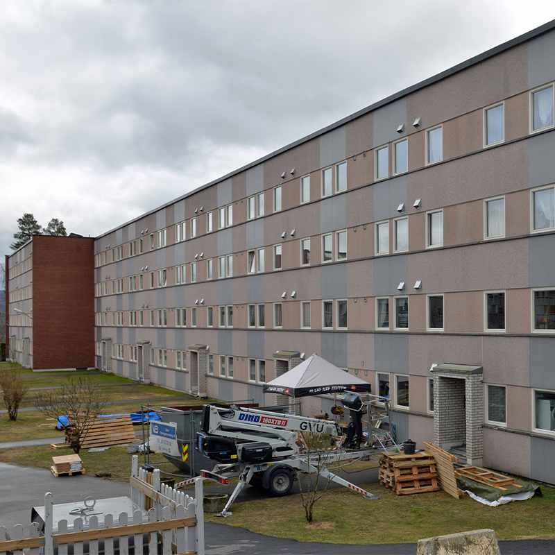 Lang blokk med grå-rosa paneler, arbeidslift, paller og telt i bakgård under overskyet himmel