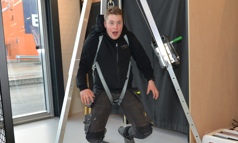 Håndverker i svart hettegenser og grå sele kneiser i aluminiumsramme med verktøy i beltet