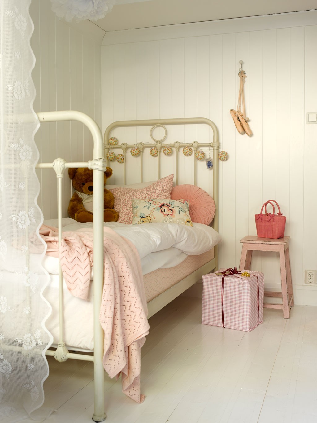 Lys barnerom med kremhvit jernseng, rosa pledd, bamse, ballerinasko og gavepakke på gulvet