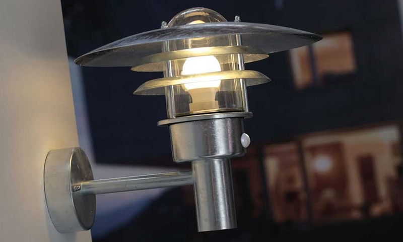 Sølvfarget vegglampe med glasskjerm og lysende pære mot mørk husfasade