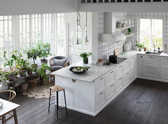 Lyst kjøkken med hvit øy, mørkt tregulv og grønne planter ved store vinduer.