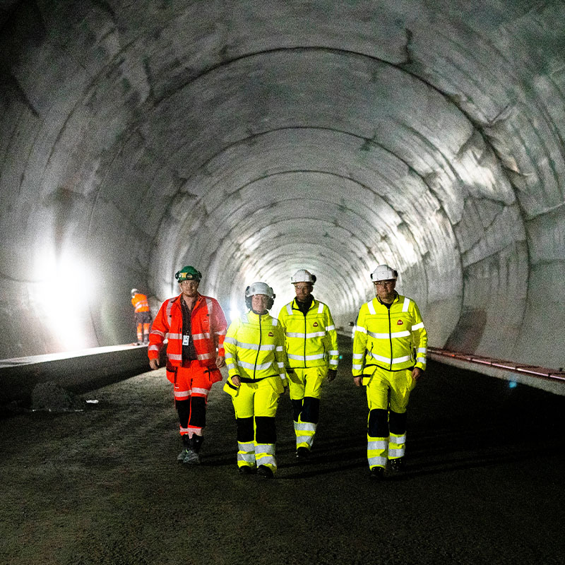 Fire anleggsarbeidere med hjelm i gule og røde refleksdresser går i betongtunnel