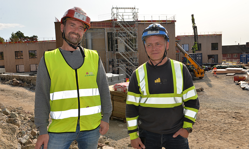 To arbeidere i gule refleksvester og hjelmer foran en uferdig bygning med stillas og kran.