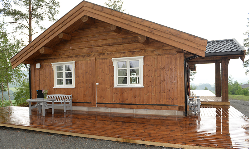 Brunt trehytte med hvite vinduskarmer, grå benk og bord på regnvåt terrasse i skog