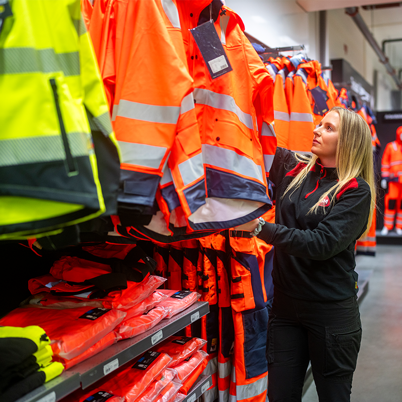 Ansatt plukker oransje arbeidsjakke med reflekser fra klesstativ fylt med verneklær.