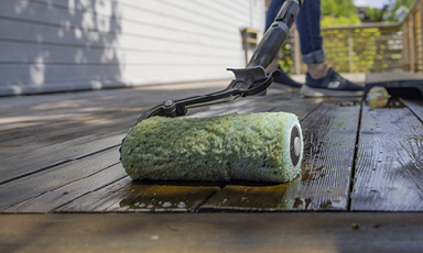 Nærbilde av grønn malerull som påfører brun beis på treterrasse, jeans og joggesko bak.