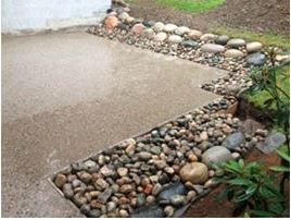 Grå betongflate med kant av flerfargede glatte elvesteiner ved grønt hagebed.