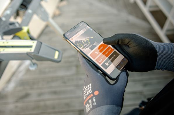 To hanskekledde hender holder smarttelefon med kart og fargede appknapper over grått trebord.