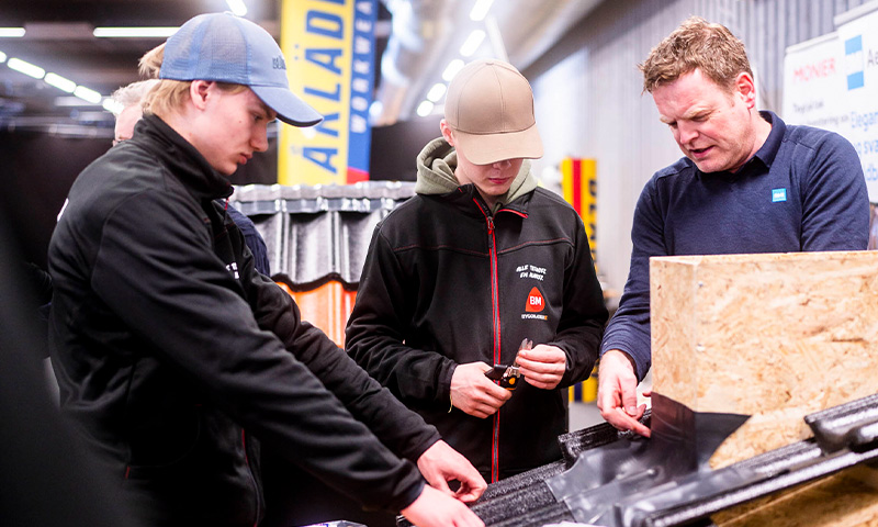 Tre håndverkere i mørke jakker måler sort takpanel i butikkmiljø.