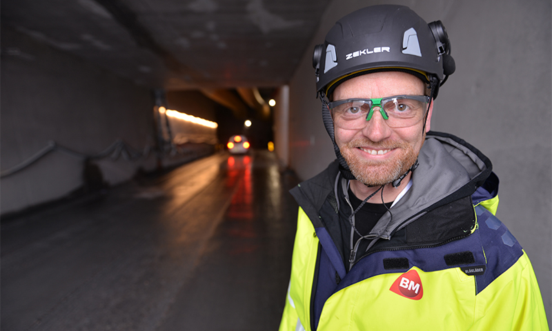 Anleggsarbeider i gul refleksjakke med rødt BM-merke og hjelm ved tunnelåpning med billys