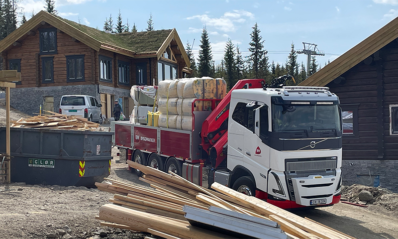 Hvit-rød lastebil med byggevarer og planker ved brune tømmerhytter under bygging i skoglandskap