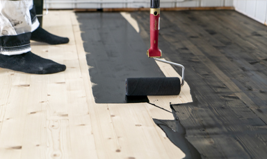 Malerulle påfører svart maling på lyst tregulv under renovering.