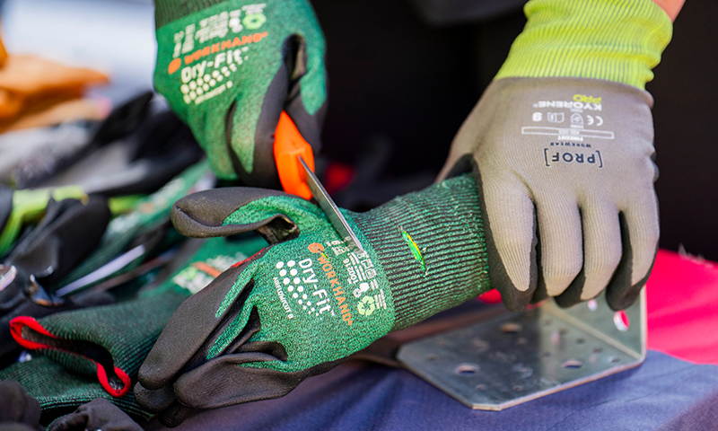 Nærbilde av hender i grønne og grå hansker som skjærer med oransje kniv ved metallbeslag.