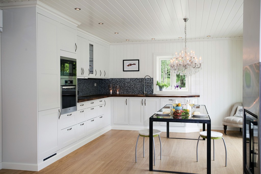 Lyst kjøkken med hvite skap, mørk mosaikk, glassbord med frukt, stoler og lysekrone.