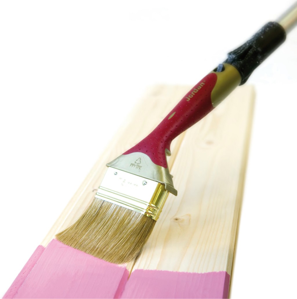 Rød-gul malekost maler en lys treplanke rosa mot hvit bakgrunn.