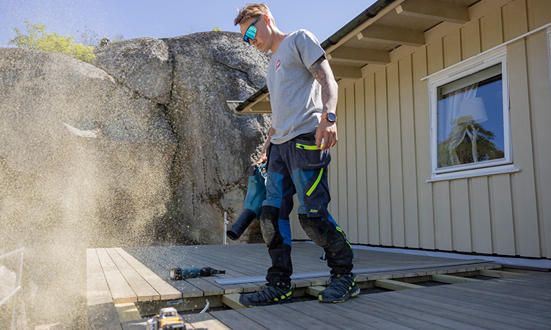 Håndverker blåser støv med varmluftpistol på uferdig terrasse ved lyst hus og granittvegg