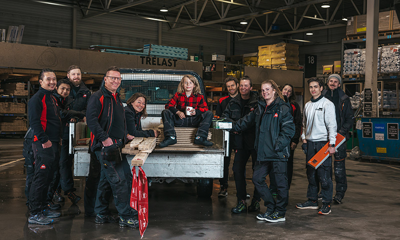 Ti håndverkere i mørke klær står ved en bilhenger med trelast i lagerhall, én i rød rutet skjorte