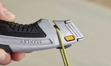 Hånd holder grå Stanley-kniv med gul knapp og kutter grønn-gul elektrisk ledning.