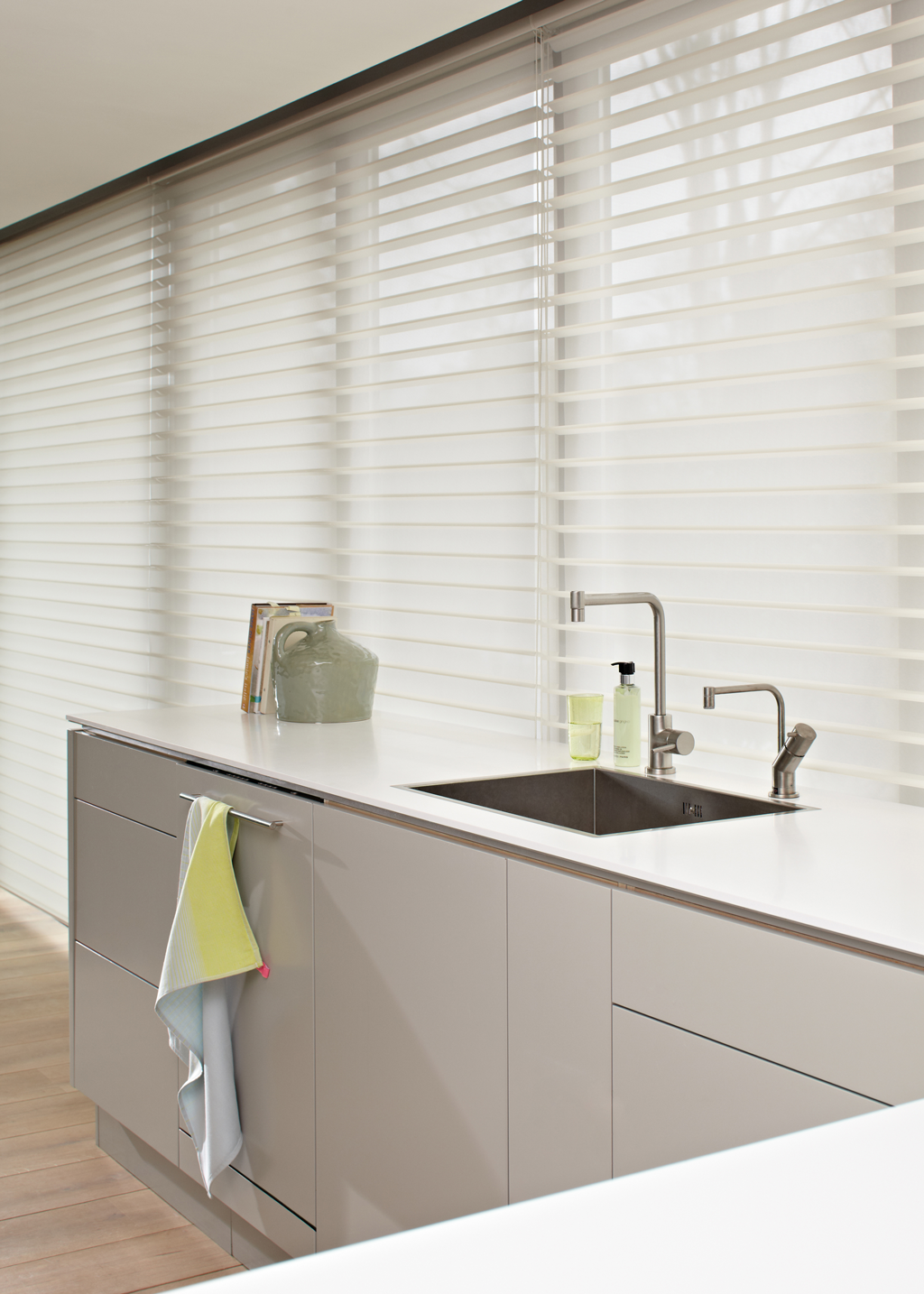 Minimalistisk kjøkken med hvit benkeplate, stålvask, pastellhåndkle og hvite lamellgardiner