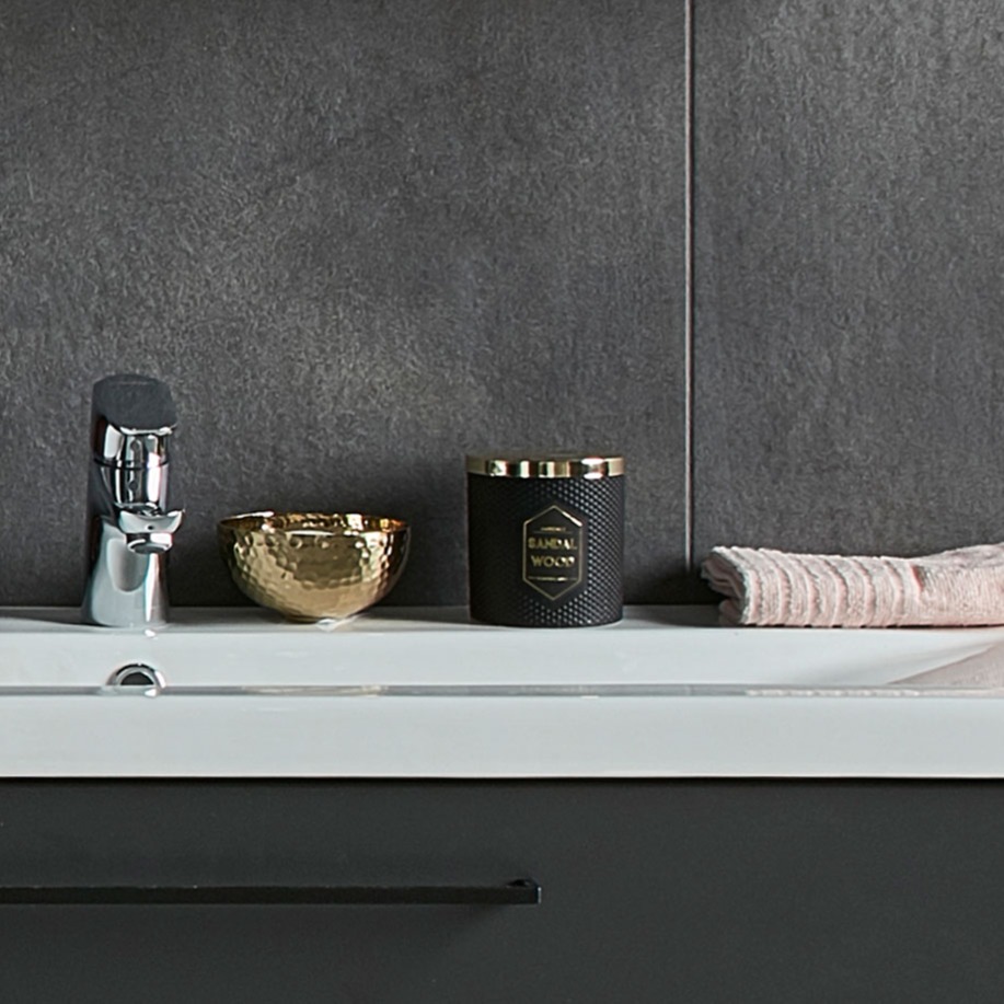 Hvitt servantbord med kromkran, gullskål, svart-gull duftlys og rosa håndkle foran grå fliser.