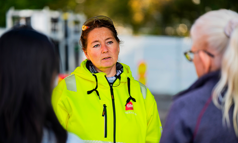 Person i neongul sikkerhetsjakke med BM-logo snakker med to personer på byggeplass.