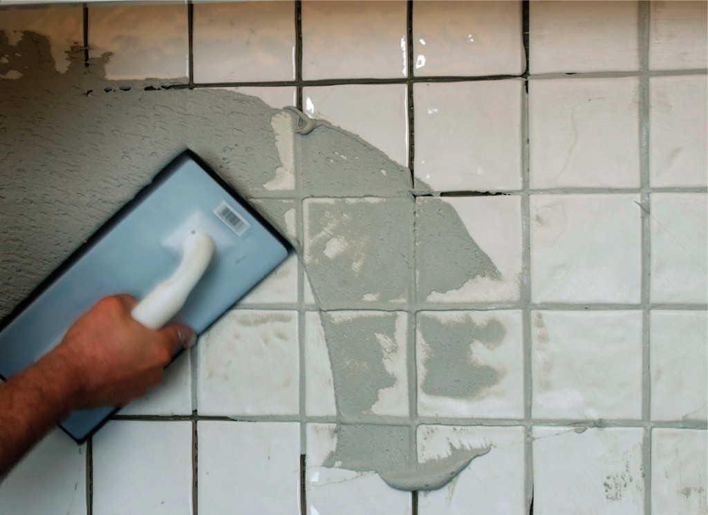 Nærbilde av hånd som med fugepresse sprer grå fugemasse på hvite fliser.