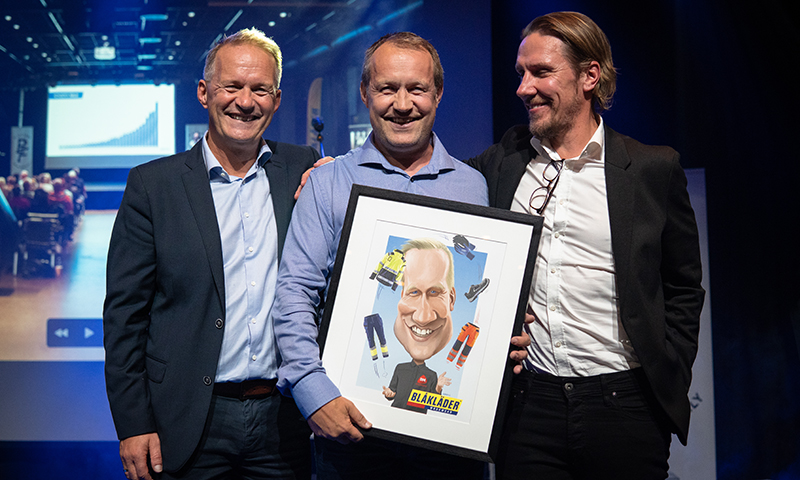 Tre menn i dresser på scene holder Blåkläder-plakat i ramme foran stor skjerm med graf.