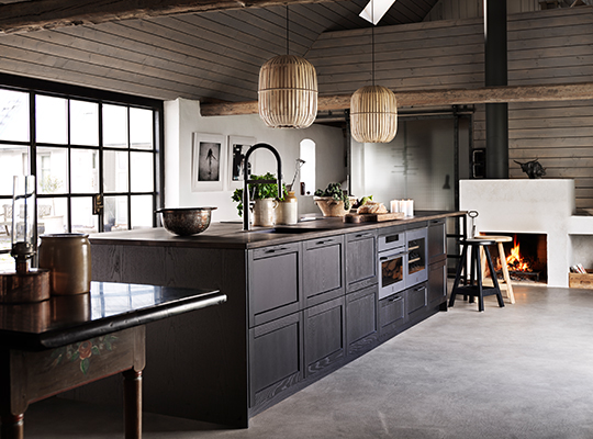 Romslig kjøkken med grå øy, rottingpendler, store vinduer og peis i tømmerloft.