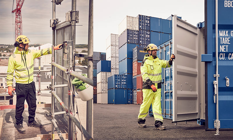 Anleggsarbeider i gul refleksjakke ved stillas og blå fraktcontainere i havneområde.