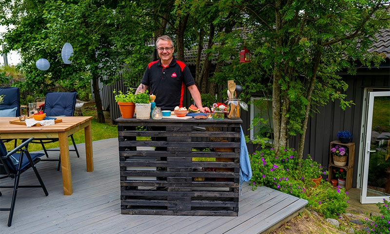 Ute på grå terrasse: svart pallbar med grønne urter, vannmelon, fat og hage i bakgrunnen