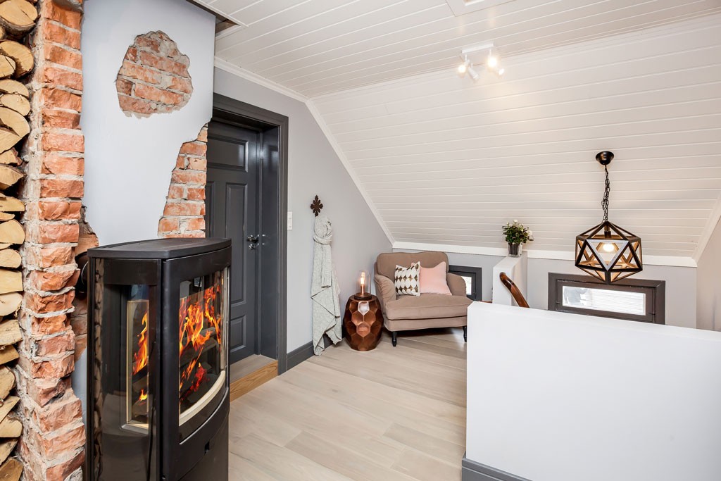 Lyst loftsrom med svart vedovn foran eksponert murstein, beige lenestol med rosa pute.