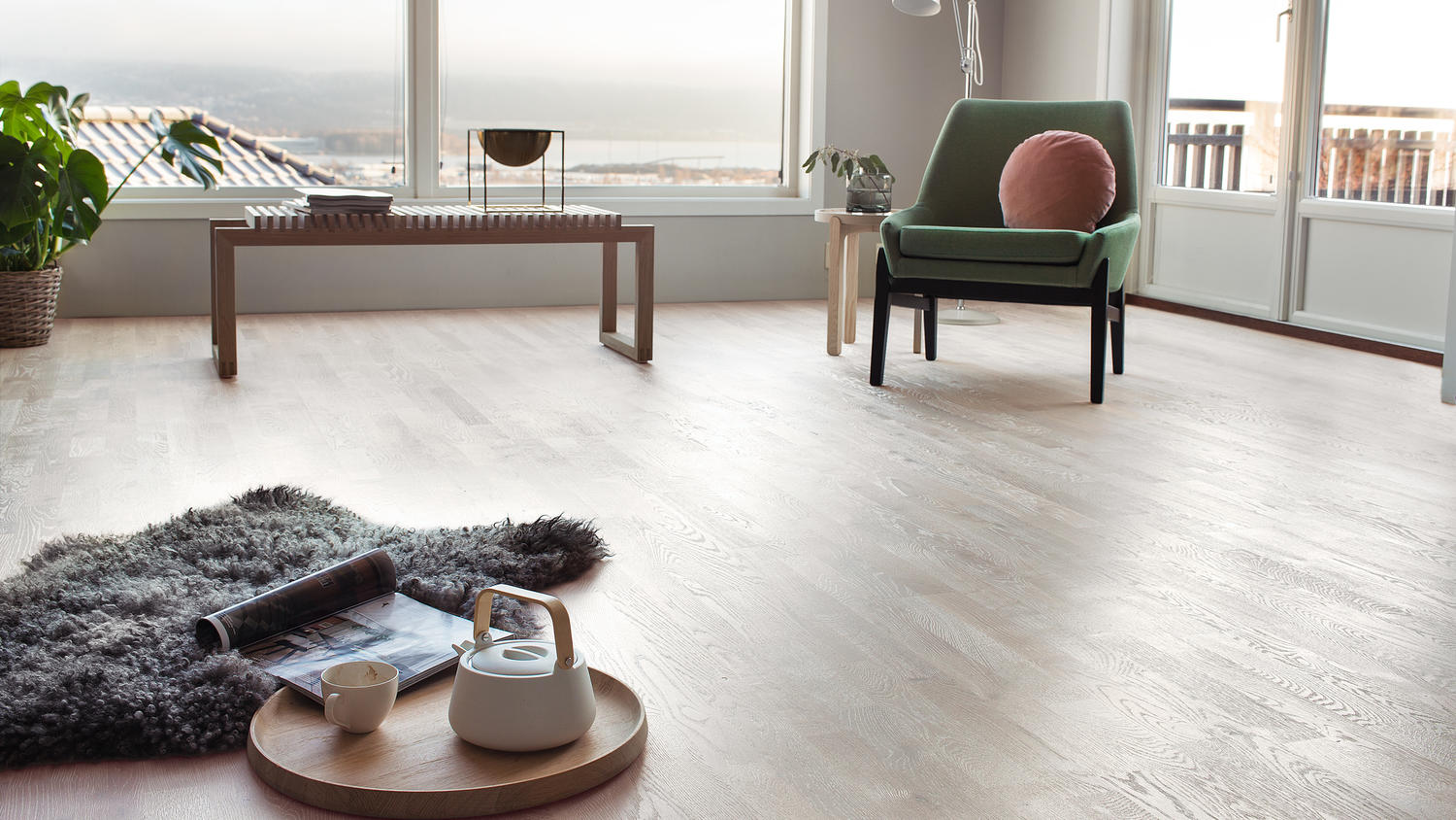 Lyst oppholdsrom med store vinduer, grønn lenestol med rosa pute, tekanne på bjørkebrett.