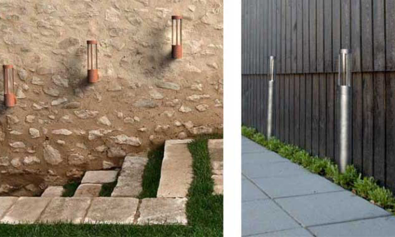 Terrakottalys på steinvegg over trapper med gress og stållys langs belegningsstein ved mørk trevegg