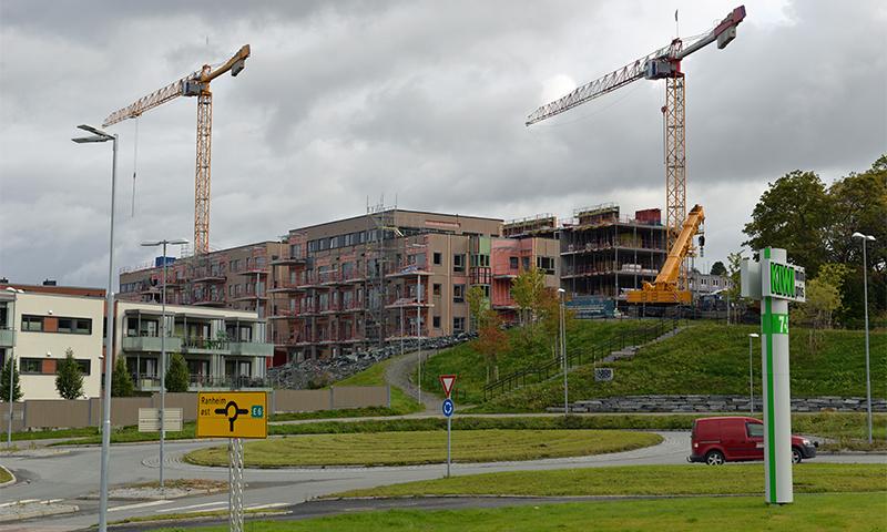 Byggeplass med gule og røde kraner over uferdige leiligheter, veiskilt, grønt gress og grå himmel