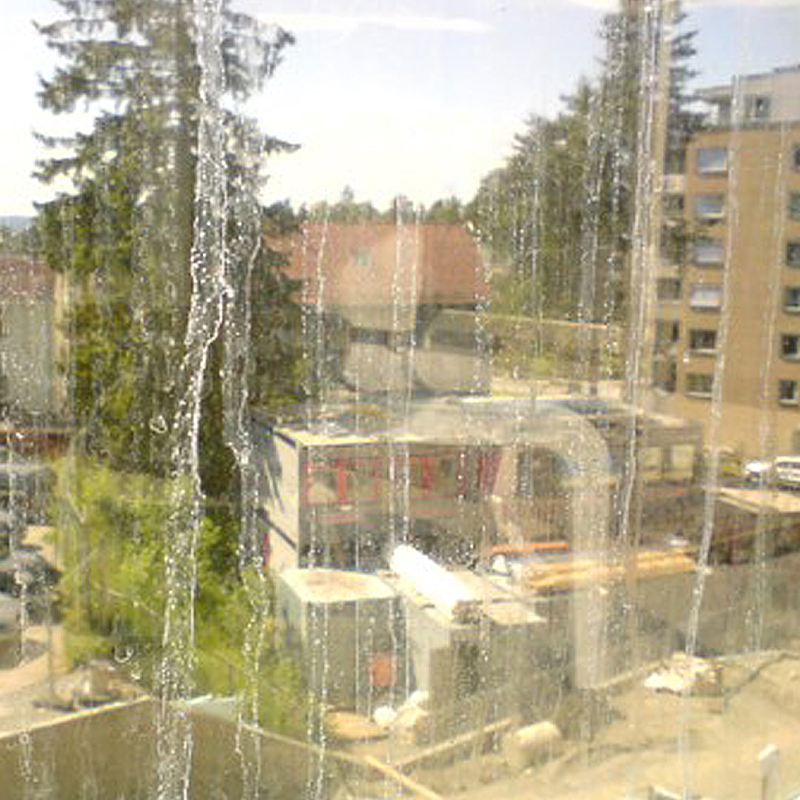 Utsikt gjennom skittent vindu mot solfylt byggeplass med hvite rør, rødt tak, grantrær