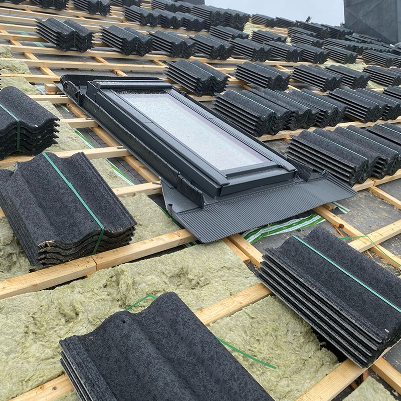 Takunderlag med trelekter, grønn isolasjon, stabler av mørkegrå takstein og takvindu