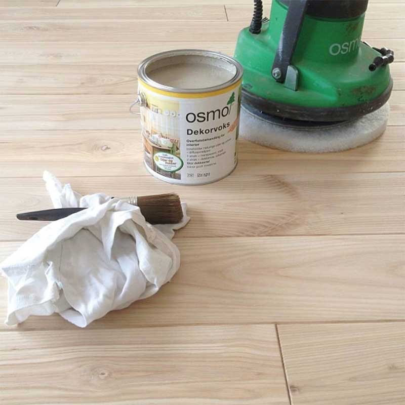 Åpen boks med Osmo dekorvoks, grønn poleringsmaskin og pensel i hvit klut på lyst tregulv.
