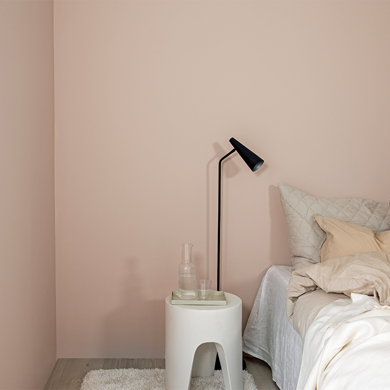 Hjørne av soverom med lyserosa vegg, hvitt bord med karaffel, svart gulvlampe og lyst sengetøy.