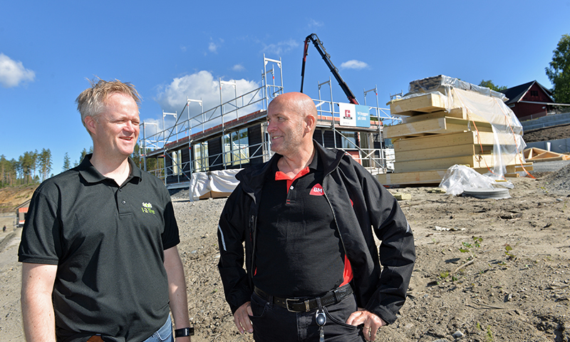 To menn i svart arbeidsklær foran nybygg med stillas, trekledningsstabler og blå himmel.