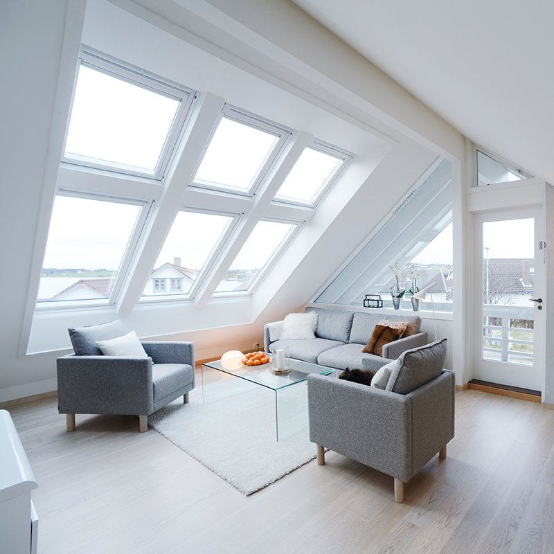 Lyst loftstue med skråtaksvinduer, sjøutsikt, grå sittegruppe og glassbord med appelsiner.