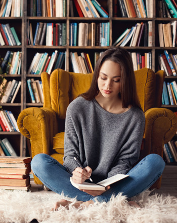 A young female author writing in her journal while sitting in front of some fully stacked bookshelves.